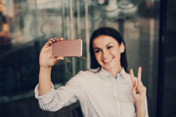Nice and beautiful girl is taking selfie and posing on camera. She is showing the piece symbol. Woman looks cheerful. She is standing at glass wall of restaurant