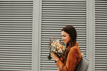 Calm and peaceful girl is standing and holding flowers. She is smelling them. Woman wears brown jacket and grey bag. Isolated on striped and white background