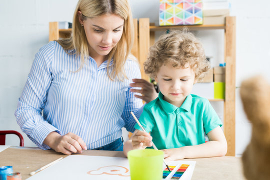 Little boy drawing picture with paint with teacher helping him in art lesson