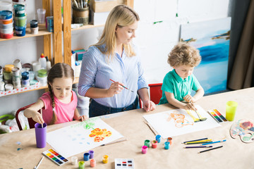 Fototapeta premium Children drawing a picture with paints and young teacher assisting them