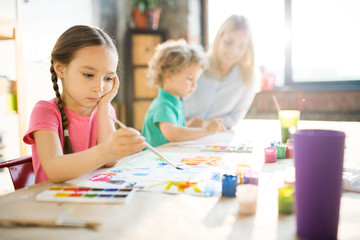 Fototapeta premium Concentrated little girl sitting at the table and painting picture with young woman and boy in the background
