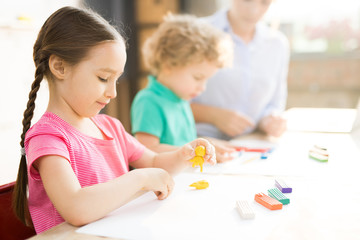 Little girl sitting at the table and play dough and mold from plasticine in art school