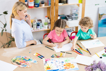 Fototapeta premium Children sitting at wooden table and drawing a picture with colorful pencils with teacher talking on cellphone and looking at how her pupils work