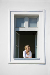 Beautiful blonde woman looks out from open window, white wall and white window frame