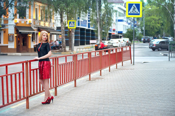 A blond girl in a red skirt and a black blouse stands on a city street in the evening. City fashion.