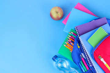 Many multi-colored school supplies falling out of a blue school backpack on a bright blue...