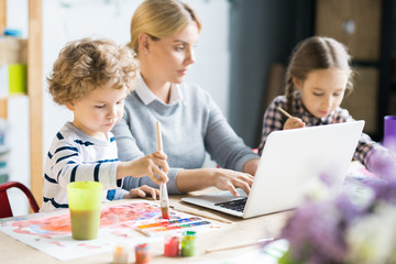 Mother helping her children with homework. She is using her laptop while kids painting a picture at the table