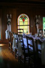 Fototapeta na wymiar Intimate Wedding Reception Table Setting with White Table Cloth in a Wood Panel Restaurant Room with Stained Glass Window and Chandelier
