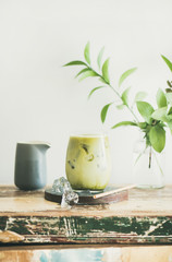 Iced matcha latte drink in glass, white wall and plant branches at background, copy space. Summer refreshing beverage cold drink