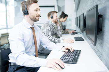 Portrait of handsome bearded man wearing headset  working with group of help desk operators sitting...