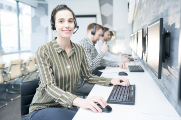 Portrait of smiling young woman  wearing headset  looking at camera while working with group of help desk operators sitting in row, copy space