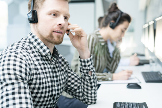 Portrait of  young man wearing headset working with group of help desk operators sitting in row and  talking to clients , copy space