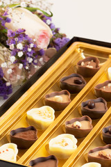 Bouquet of pink roses with baby breath and purple flowers next to fancy box of chocolates.