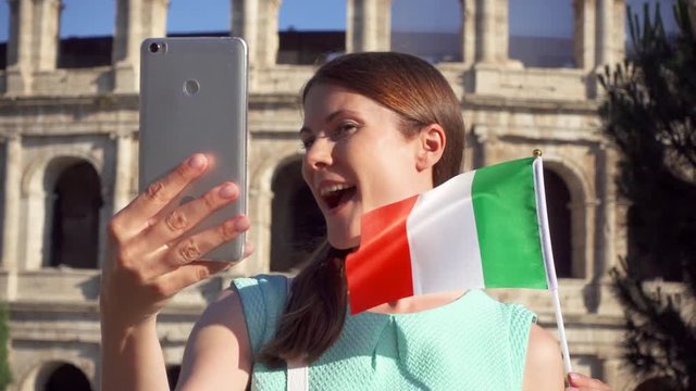 Woman doing selfie on mobile near famous attraction Colosseum in Rome, Italy. Teenage girl waving Italian flag in slow motion. Female tourist enjoying european vacation. Student travel through Europe