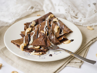 Thin chocolate pancakes with condensed milk, chocolate sauce and cashew on a white plate. Stack of crepes, russian blin. Tasty breakfast. Close up, selective focus