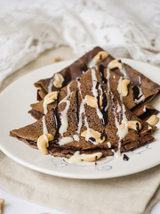Thin chocolate pancakes with condensed milk, chocolate sauce and cashew on a white plate. Stack of crepes, russian blin. Tasty breakfast. Close up, selective focus