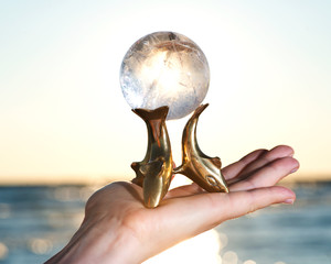 Woman's hand holding Lemurian Clear Quartz Sphere on Dolphins Brass Stand at the sunrise in front of the lake.