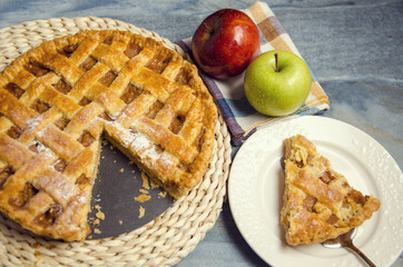 Slice of apple pie on plate next to whole pie and some apples 