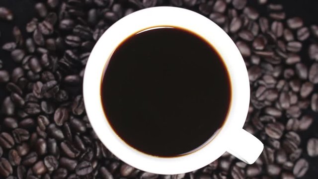 Black coffee in white cup with coffee beans rotating on dark surface.