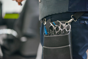 Close up of barber's leather bag with metallic sharp scissors hanging on waist. Hairstyler wearing...
