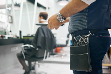 Close up of barber's leather bag with metallic sharp scissors for haircutting. Hairstyler wearing dark blue trousers, grey waistcoat, handwatch. Keeping black plastic comb. Client sitting behind.