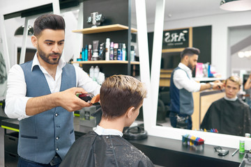 Doing new modern hairstyle for young man in barber shop. Professional hairstylist using clipper, looking concentrated. Male model sitting in front of big mirror, covered with special black cape.