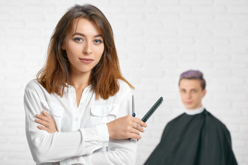 Woman hairstylist looking at camera holdingblack plastic comb and sharp metallic scissors. Looking at camera wearing stylish white shirt. Young client with toned lilac hair sitting behind.