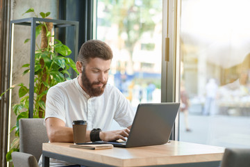 Fototapeta premium Young man working with laptop sitting near panoramic windows. Drinking coffee gron paper can, wearing casual white shirt, leather bracelet, having brown hair and beard, looking concentrated.