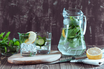 Fresh cold water with lemon, cucumber, mint and ice in a pitcher and glass on a table.