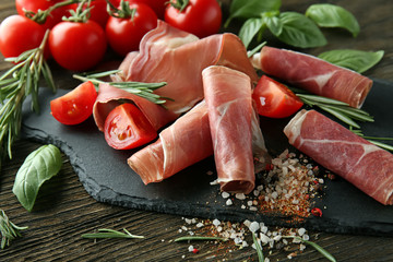 Slate board with tasty prosciutto slices, tomatoes, spices and herbs on wooden table