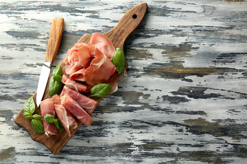 Wooden board with tasty prosciutto slices and basil on table