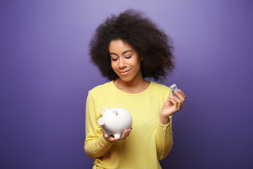 Young African-American woman with money and piggy bank on color background. Savings concept