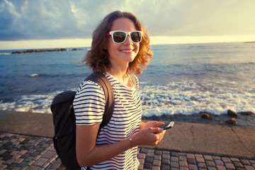 Happy casual woman walking in a port promenade and laughing watching media content on a smart phone with sea at sunset in the background