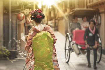 Fototapeta premium Maiko spaceruje aleją Kioto w świetle zachodu słońca przekraczając rikszę.