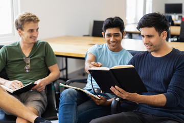 Three fellow students studying, reading textbooks and talking. Young men sitting in armchairs in classroom or library. Education concept.