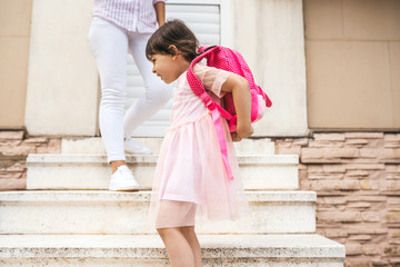 Mother standing on the stairs against the home outdoor saying goodbye to her daughter as she leave for kindergarten. Cute happy little girl wears backpack going to preschool. Education, people concept