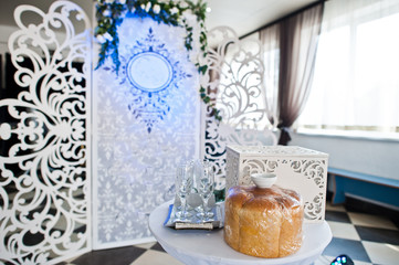 Special photo zone with small wooden wall decorated with flowers and wedding couple's initials. Table with box and bread on it.