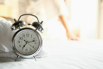 Alarm clock standing on bedside table has already rung early morning to wake up woman is stretching...