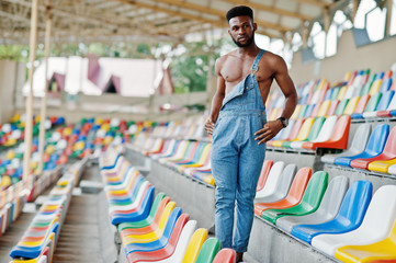 Handsome sexy african american bare torso man at jeans overalls posed on colored chairs at stadium. Fashionable black man portrait.