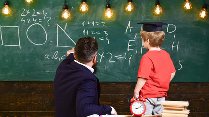 Teacher and schoolboy turned back in front of the green board. Kid in graduation cap learning shapes. Adult pointing at triangle drawn on the board.