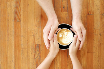 Young couple in love holding hands over black cup of coffee with swan shaped foam latte art on vintage grunged scratched table. Man & woman body language. Background, top view, close up, copy space.