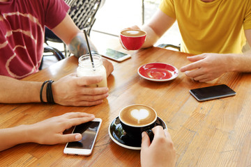 Fototapeta na wymiar Group of friends drinking cappuccino at coffee shop, staring at smartphones, checking likes & messages. Addiction to social media. Young men & women holding cell phones & drinks. Background, top view.