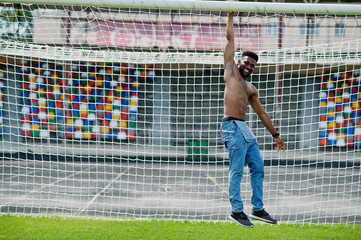 Handsome sexy african american muscular bare torso man at jeans overalls posed at green grass agasinst football gates of stadium field. Fashionable black man portrait.