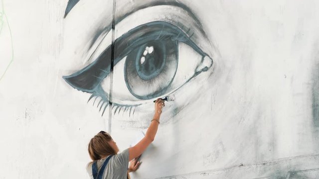 Beautiful Young Blonde Girl making graffiti of big eye with aerosol spray on urban street wall. Cinematic toned footage. Creative art. Talented student in denim overalls drawing picture. 4k