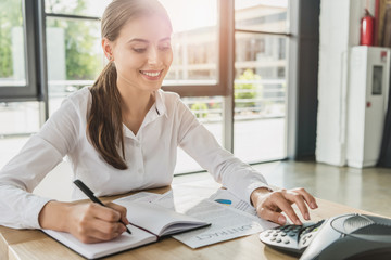 young smiling businesswoman doing paperwork and pushing button of conference phone on table at office