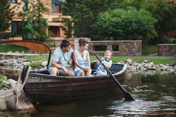Fototapeta premium Mother, father and son sit in a wooden boat with oars and feed the fish with bread.