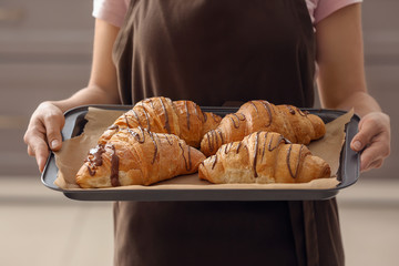Young woman holding baking tray with fresh croissants indoors