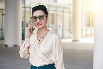 Happily smiling beautiful Asian female in white blouse and sunglasses standing outdoors and talking on mobile phone looking at camera on blurred background