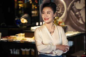 Gorgeous happy Asian female in white blouse standing with folded arms in cafe and looking away smiling on blurred background of table with food 