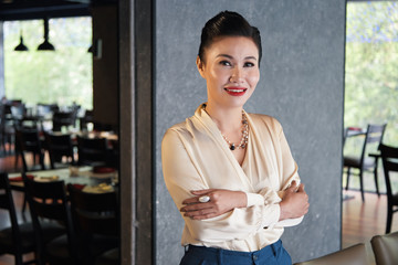Beautiful young happily smiling Asian female standing with folded arms at restaurant wall and...